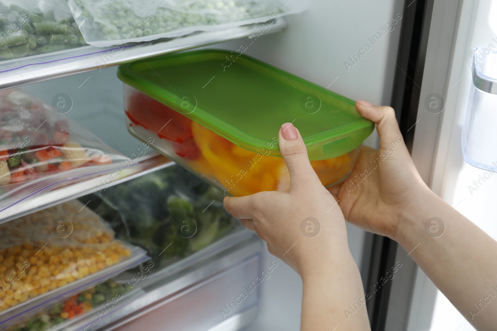 Photo of Woman taking glass container with frozen cut bell pepper from refrigerator, closeup