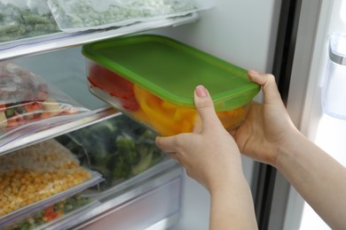 Photo of Woman taking glass container with frozen cut bell pepper from refrigerator, closeup