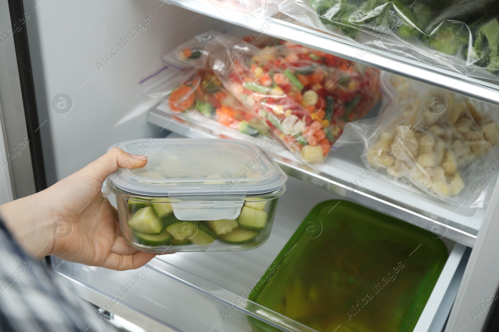 Photo of Woman taking glass container with frozen chopped zucchini from refrigerator, closeup