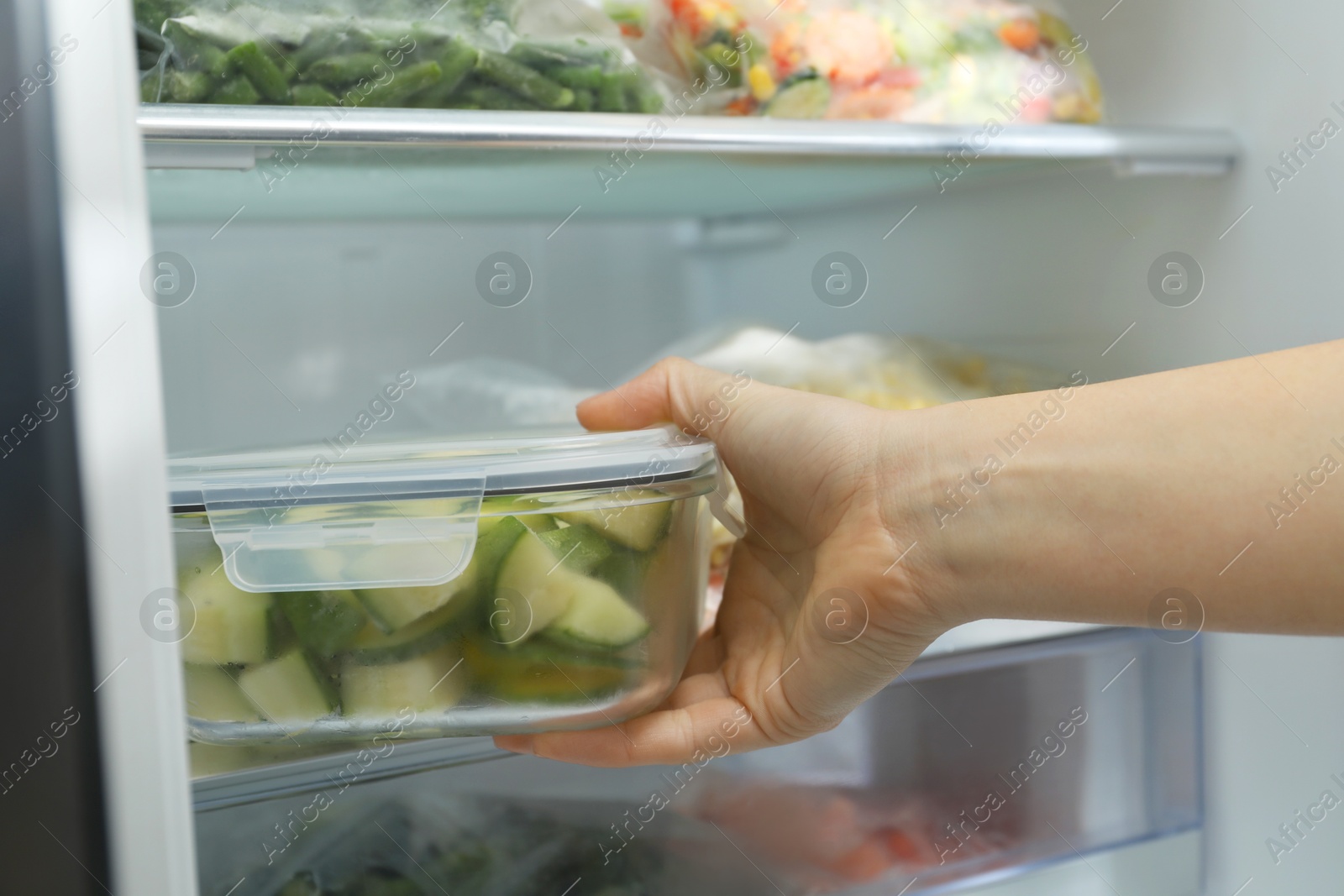 Photo of Woman taking glass container with frozen chopped zucchini from refrigerator, closeup