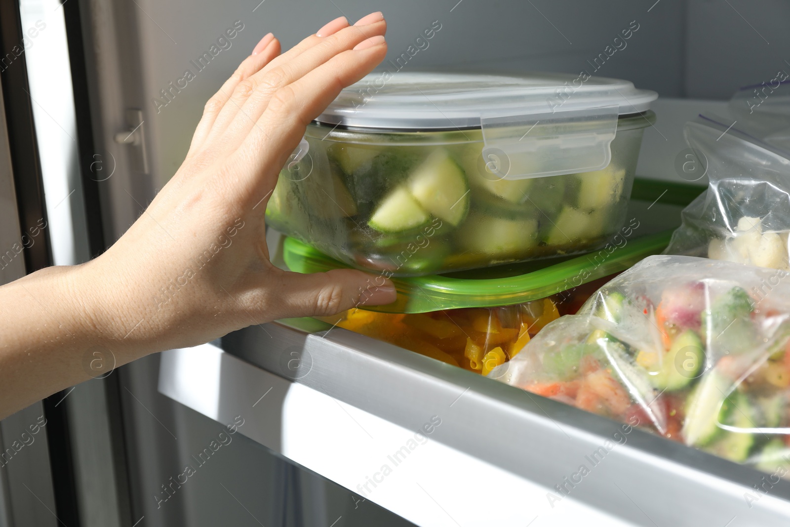 Photo of Woman taking glass container with frozen chopped zucchini from refrigerator, closeup