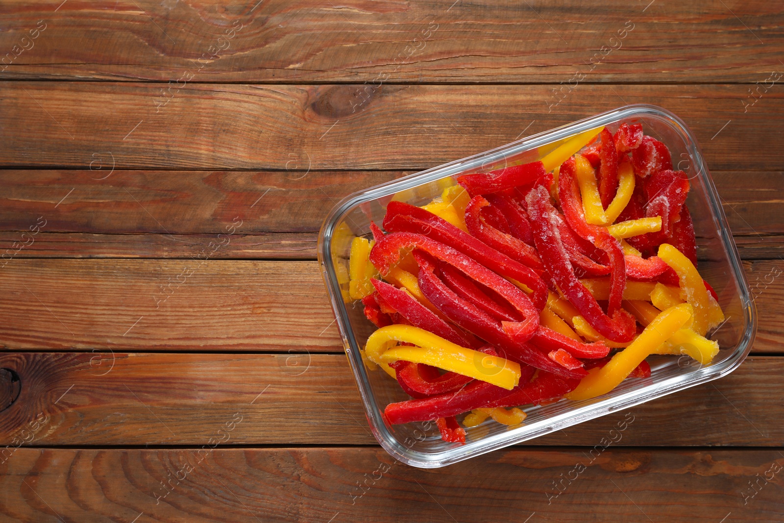 Photo of Slices of frozen bell peppers in glass container on wooden table, top view. Space for text