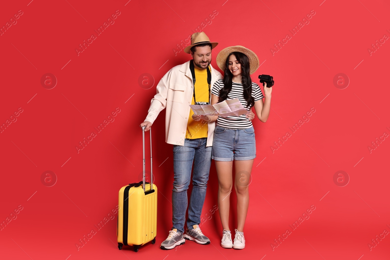 Photo of Tourism. Happy couple in hats with suitcase, map and binoculars on red background