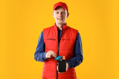 Photo of Happy courier with payment terminal and debit card on yellow background