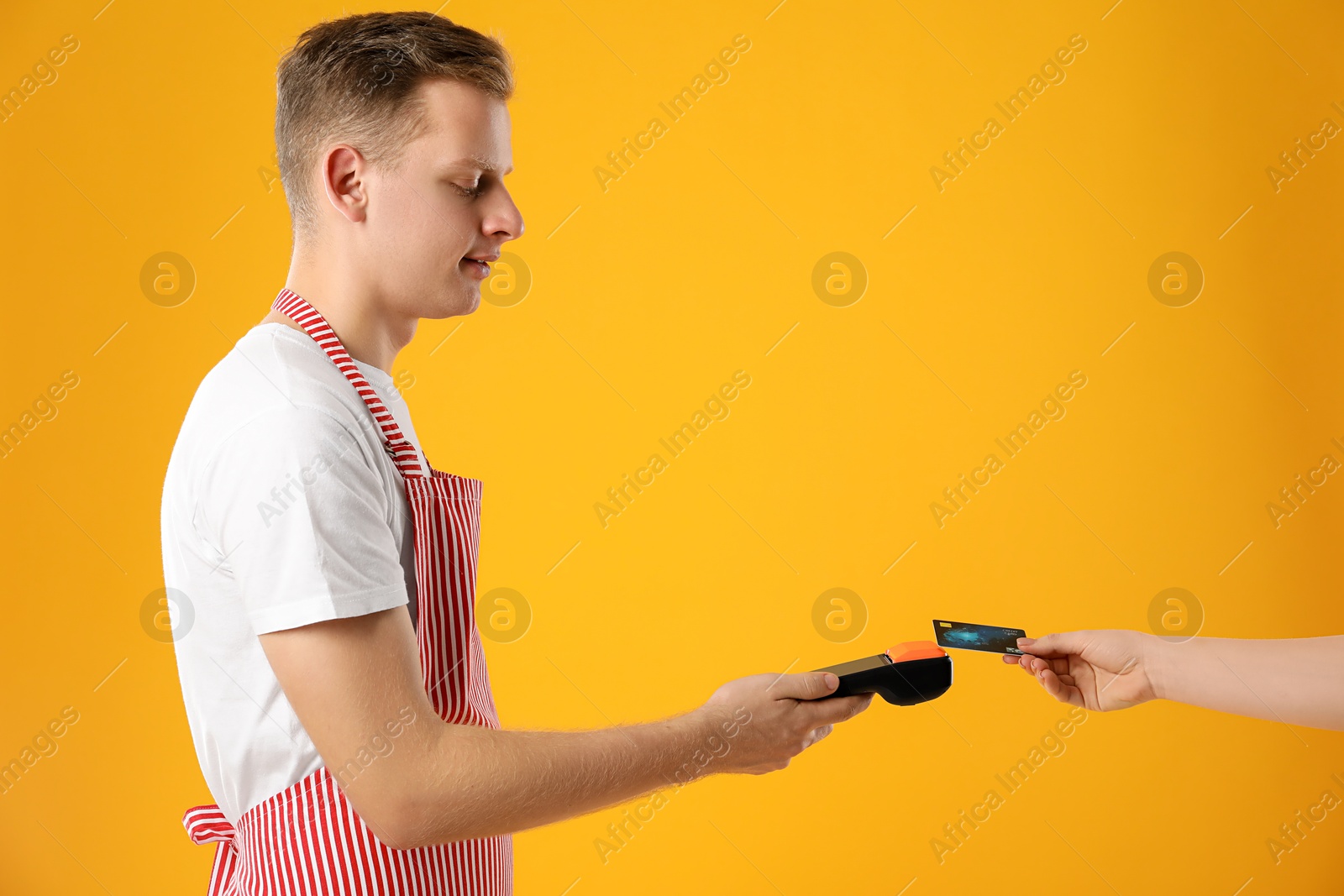 Photo of Young man in apron taking payment from client via terminal on yellow background