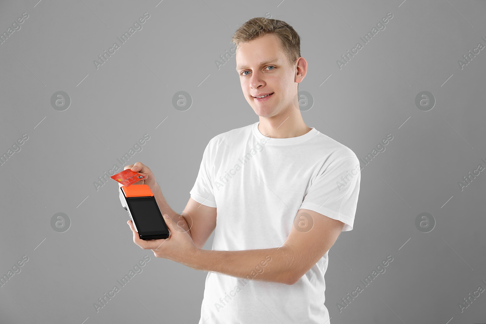 Photo of Happy young man with payment terminal and debit card on light grey background