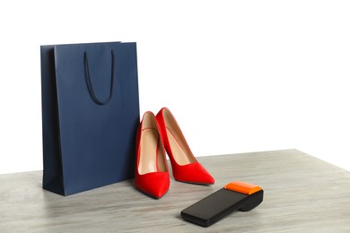 Photo of Payment terminal, shopping bag and high heeled shoes on wooden table against white background