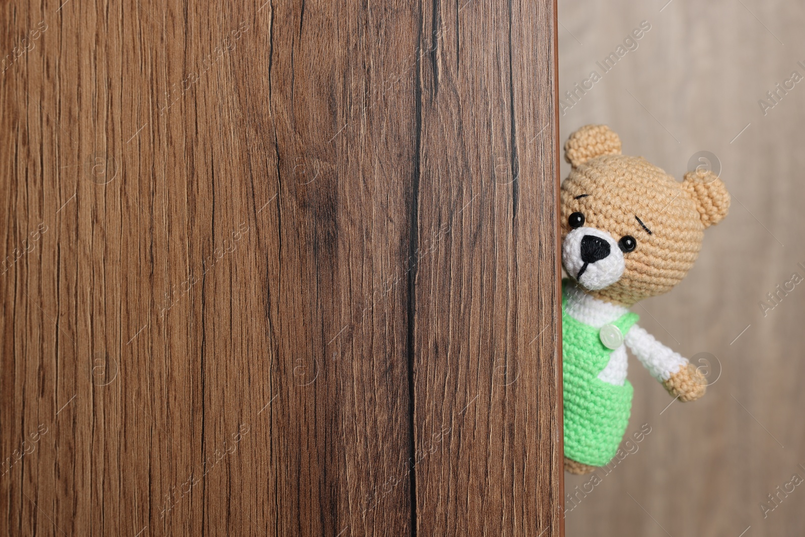 Photo of Cute knitted bear peeking out of wooden door, space for text