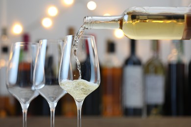 Photo of Pouring white wine into glass against blurred background with festive lights, closeup. Bokeh effect