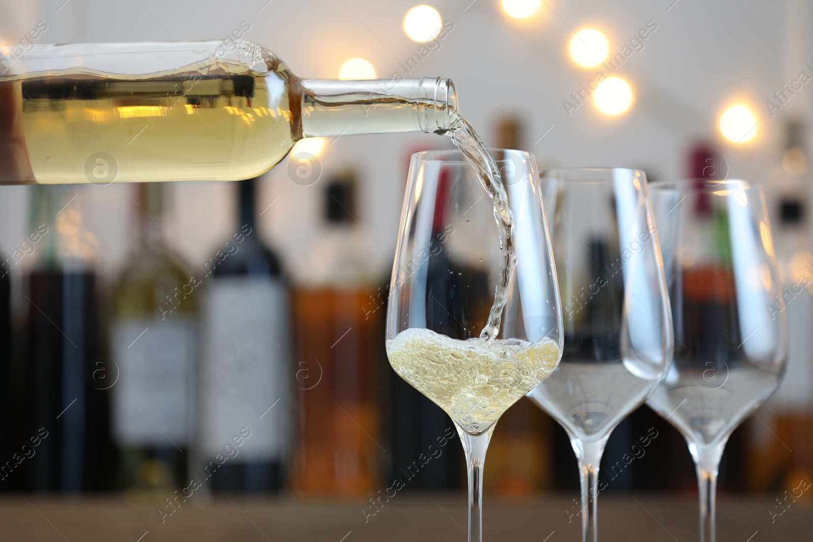 Photo of Pouring white wine into glass against blurred background with festive lights, closeup. Bokeh effect