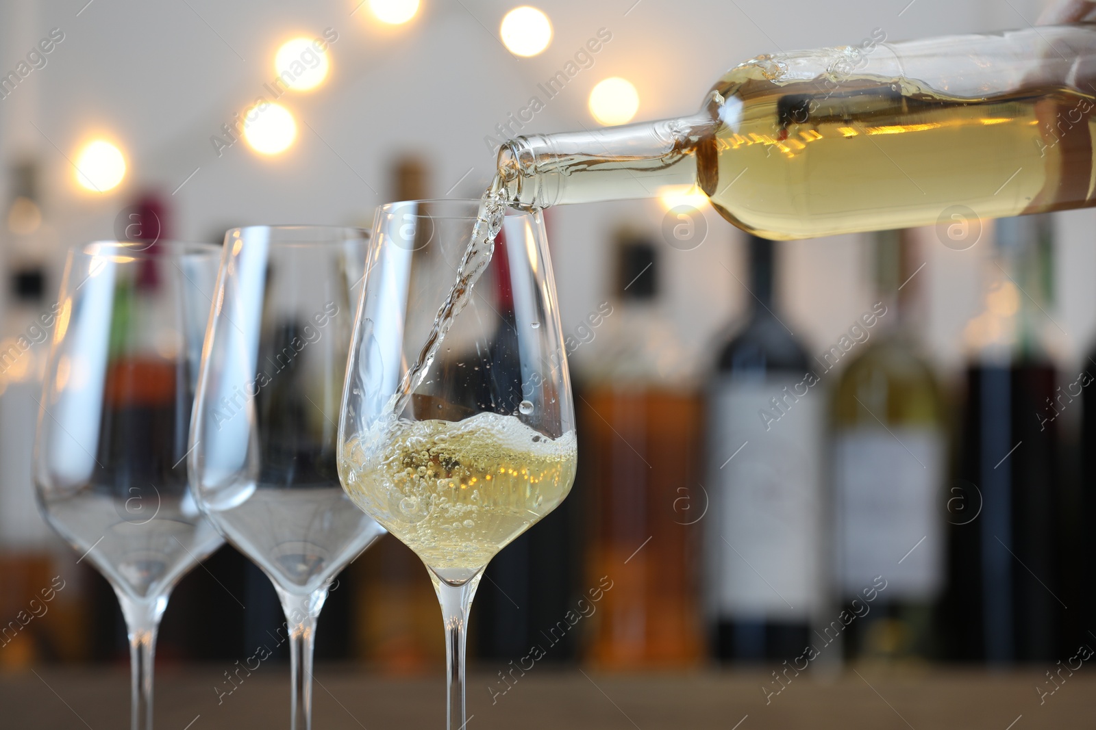 Photo of Pouring white wine into glass against blurred background with festive lights, closeup. Bokeh effect