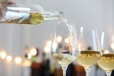 Photo of Pouring white wine into glass against blurred background with festive lights, closeup. Bokeh effect