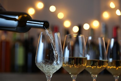 Photo of Pouring white wine into glass against blurred background with festive lights, closeup. Bokeh effect