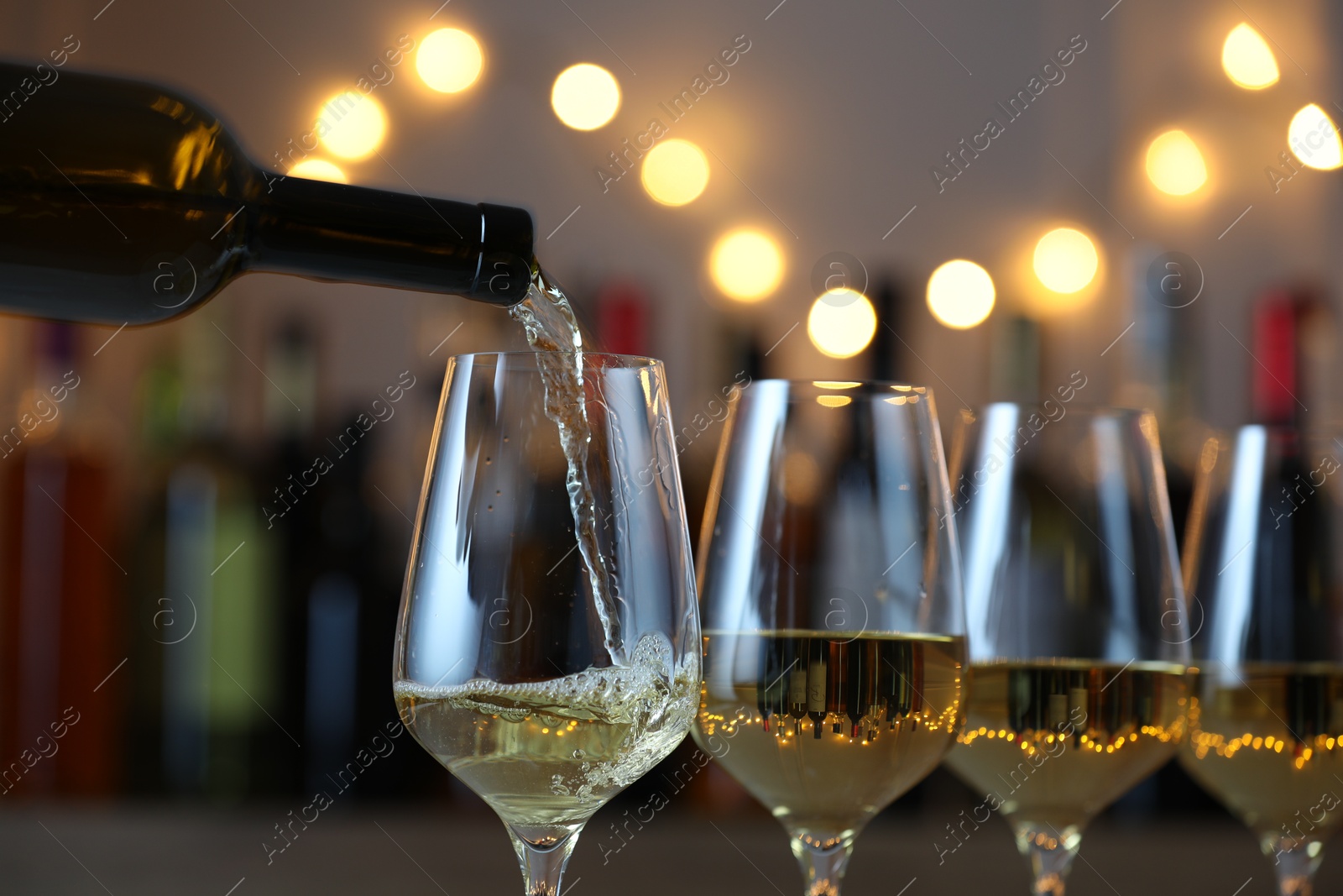 Photo of Pouring white wine into glass against blurred background with festive lights, closeup. Bokeh effect