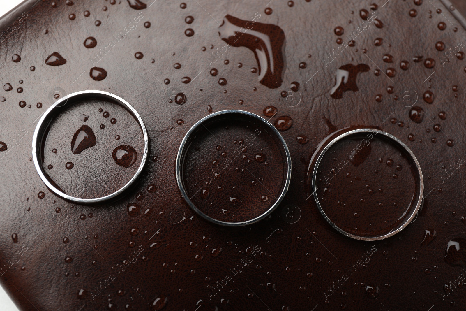 Photo of Stylish rings on leather surface with drops, flat lay