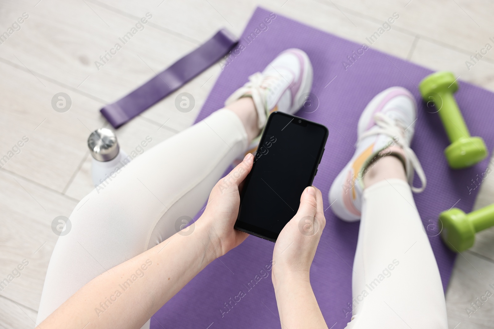 Photo of Woman with smartphone and gym equipment on floor indoors, above view