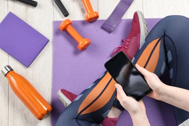 Photo of Woman with smartphone, planner and gym equipment on floor indoors, top view