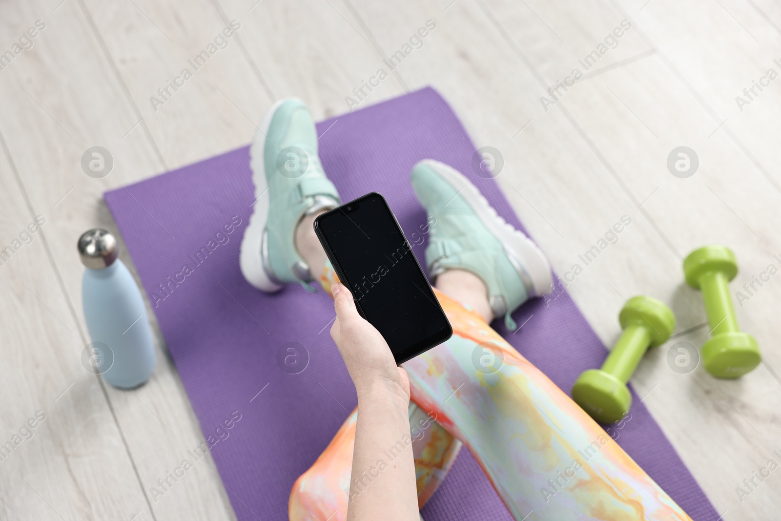 Photo of Woman with smartphone and gym equipment on floor indoors, above view