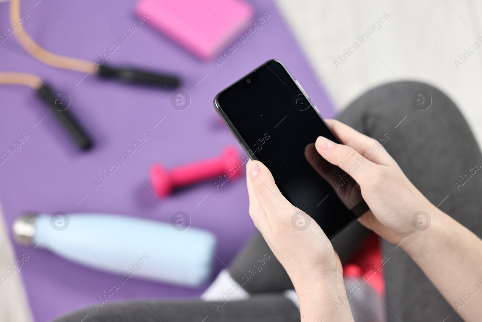 Photo of Woman with smartphone and gym equipment on floor indoors, closeup. Space for text