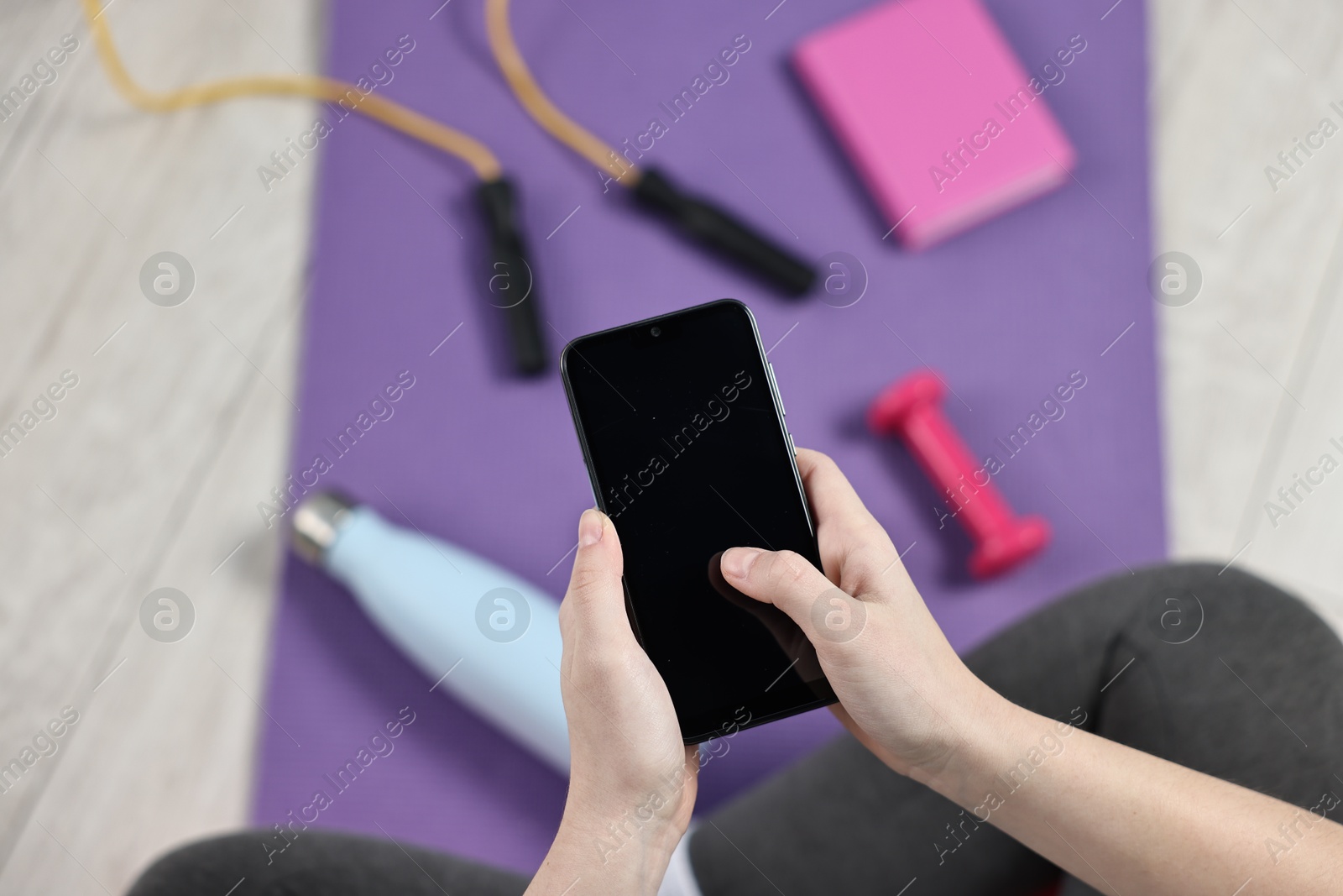 Photo of Woman with smartphone and gym equipment on floor indoors, above view