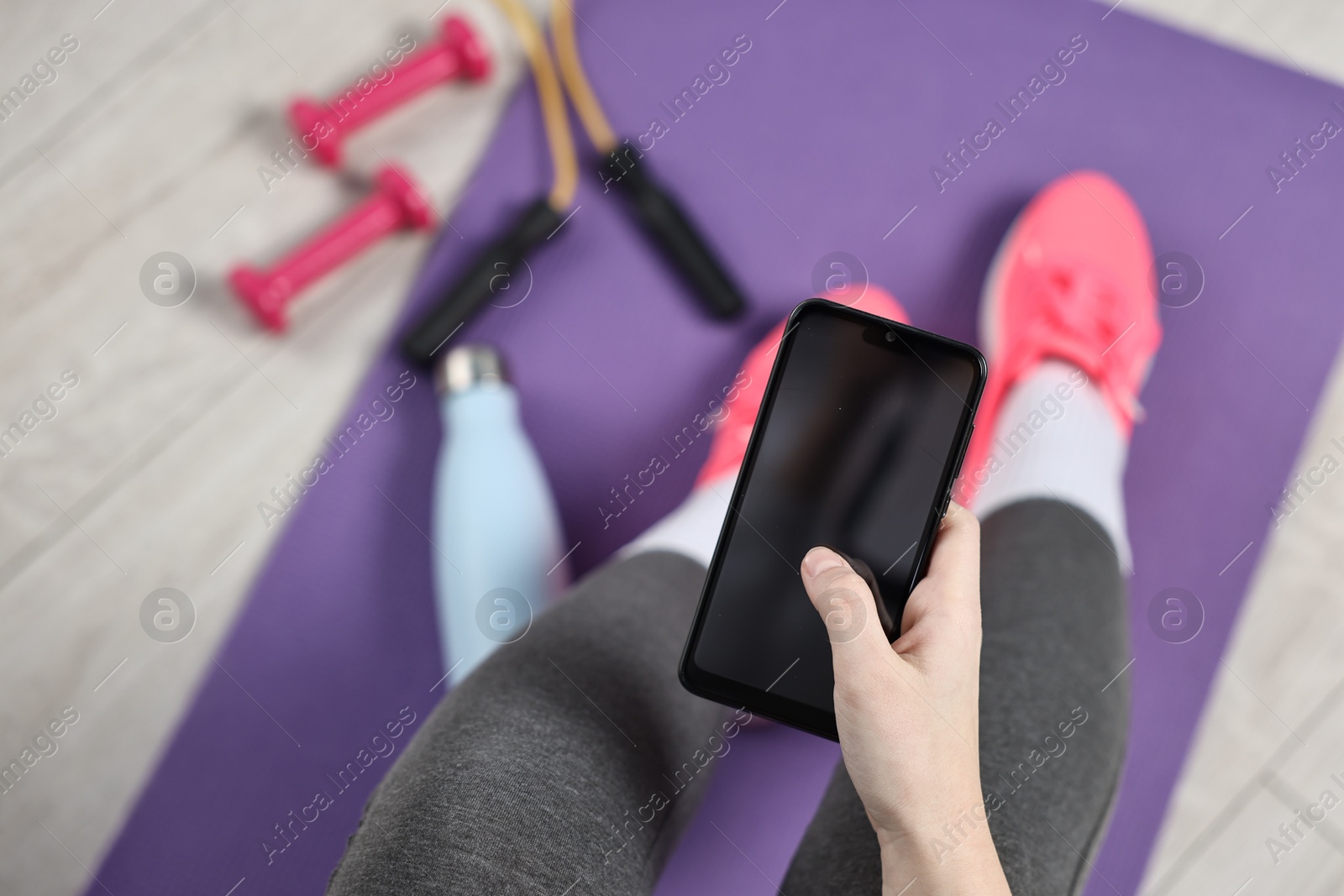 Photo of Woman with smartphone and gym equipment on floor indoors, above view