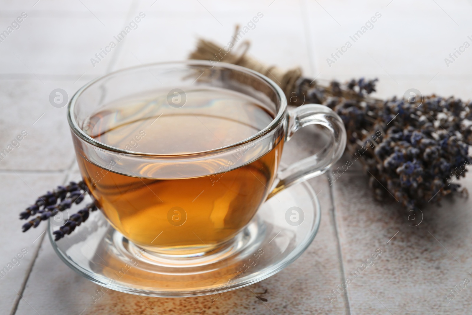 Photo of Aromatic lavender tea in glass cup and dry flowers on white tiled table, closeup