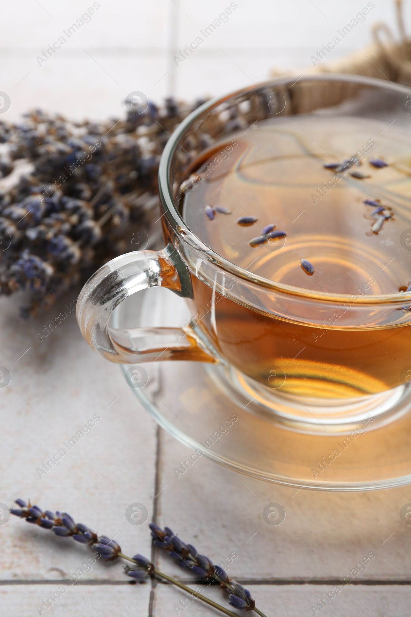 Photo of Aromatic lavender tea in glass cup and dry flowers on white tiled table, closeup