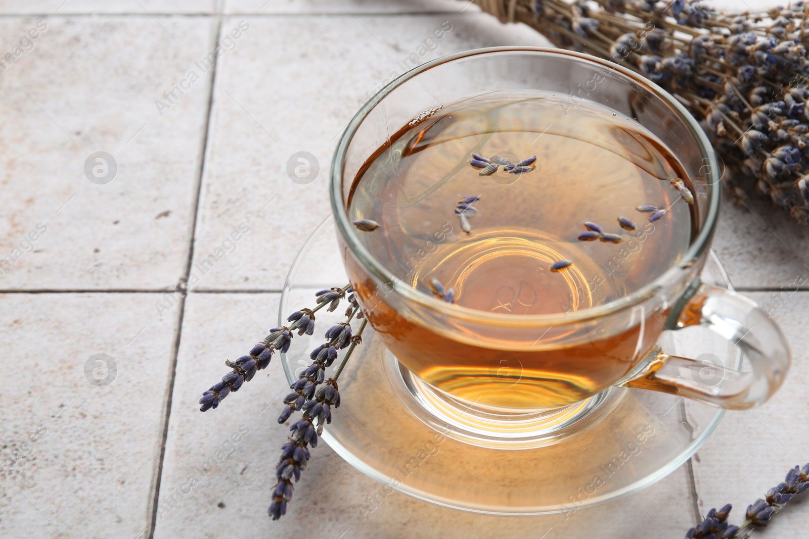 Photo of Aromatic lavender tea in glass cup and dry flowers on white tiled table, closeup. Space for text