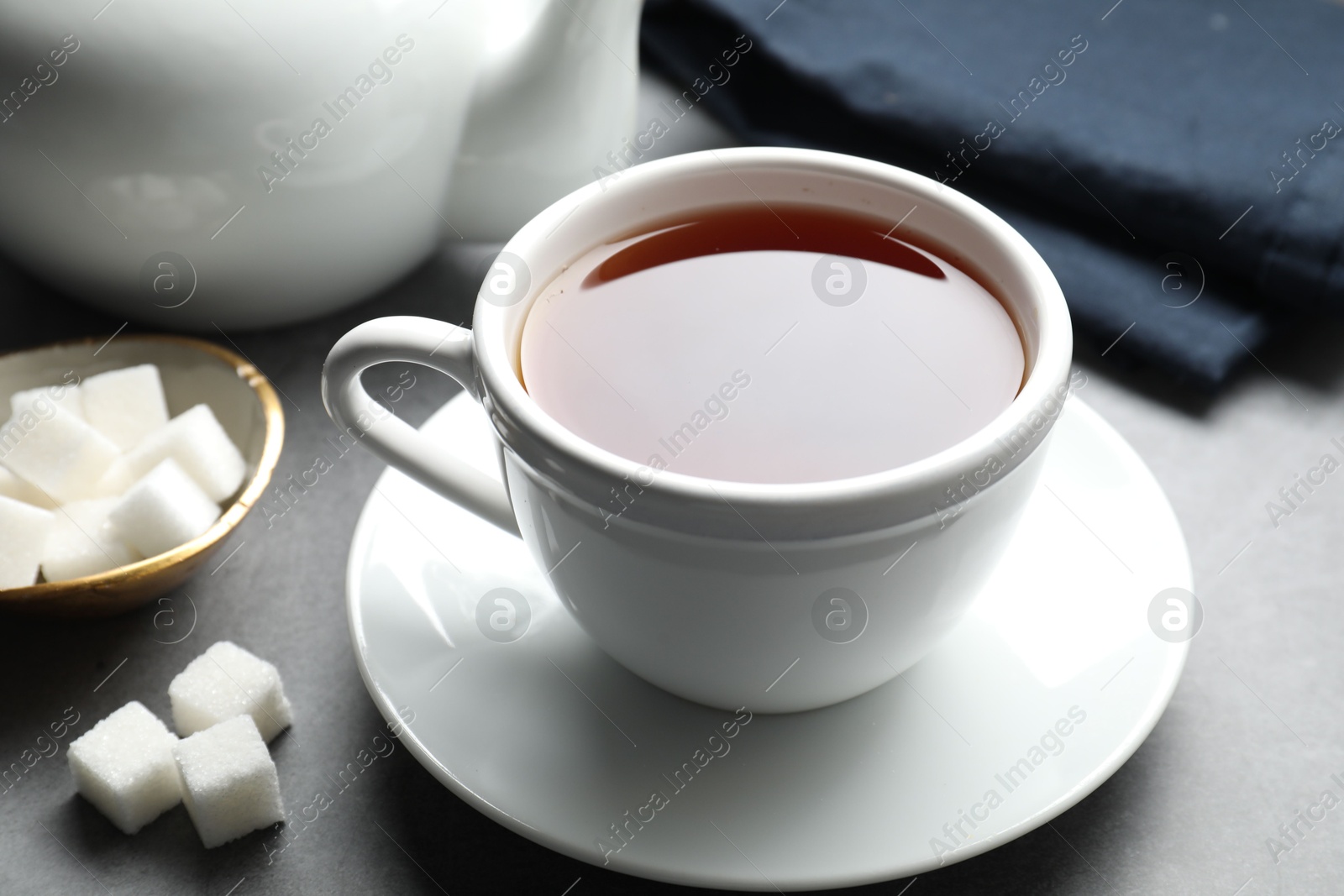 Photo of Refreshing black tea in cup and sugar cubes on grey textured table, closeup