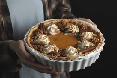 Woman holding homemade pumpkin pie with whipped cream, seeds and cinnamon on black background, closeup