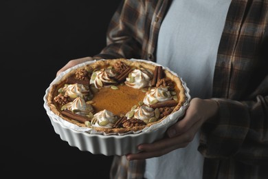 Photo of Woman holding homemade pumpkin pie with whipped cream, seeds and cinnamon on black background, closeup