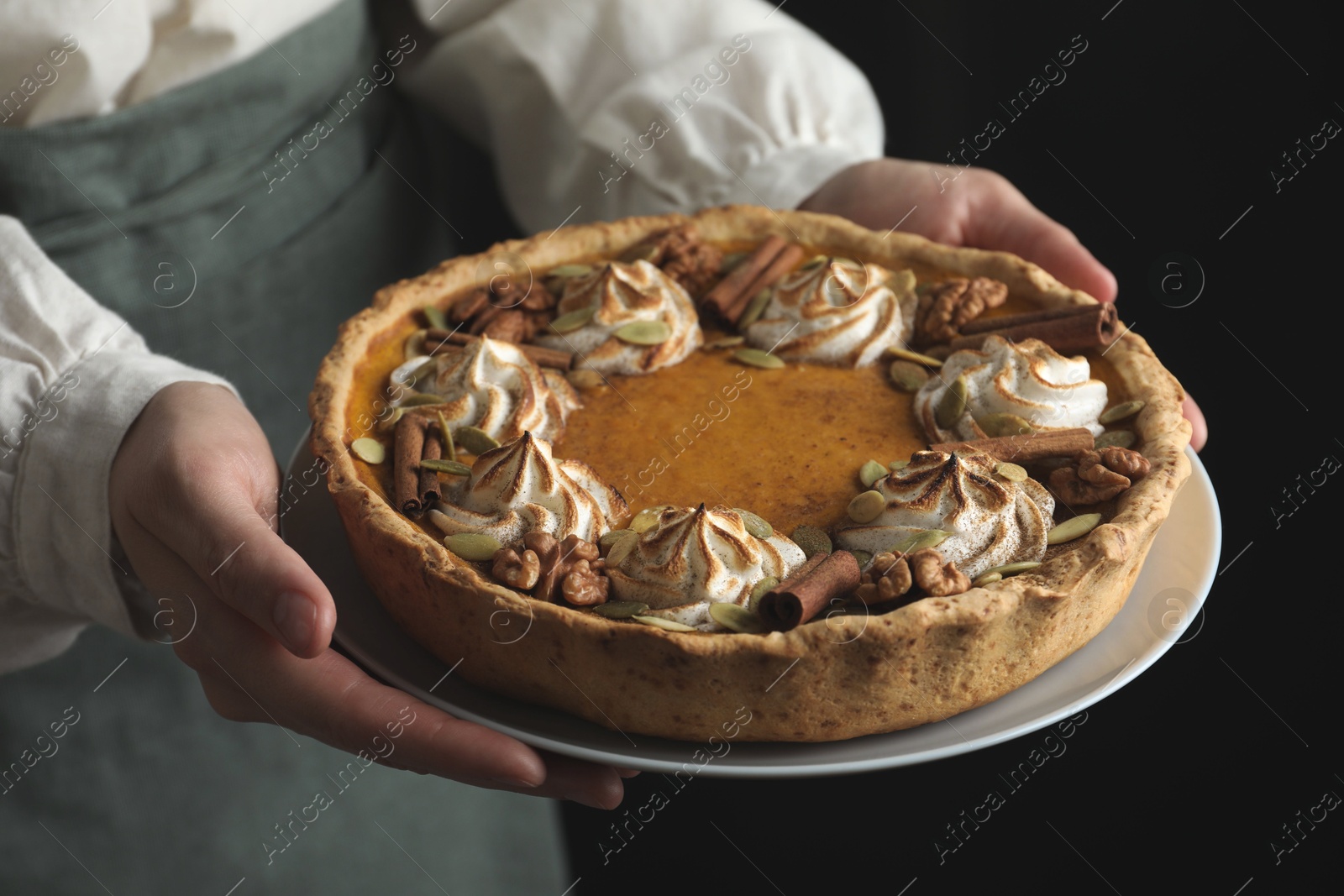 Photo of Woman holding homemade pumpkin pie with whipped cream, seeds and cinnamon on black background, closeup