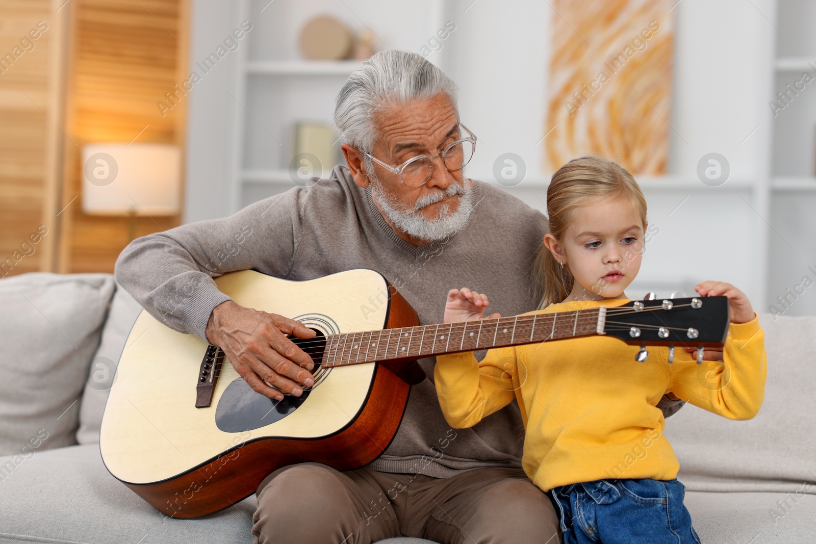 Photo of Grandpa teaching his granddaughter to play guitar on sofa at home