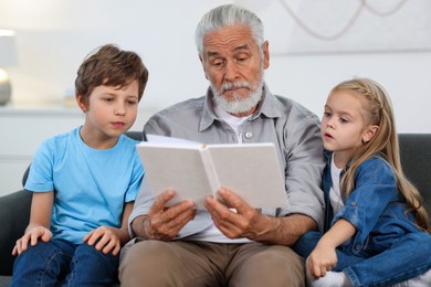 Photo of Grandpa and his grandkids reading book together on sofa at home