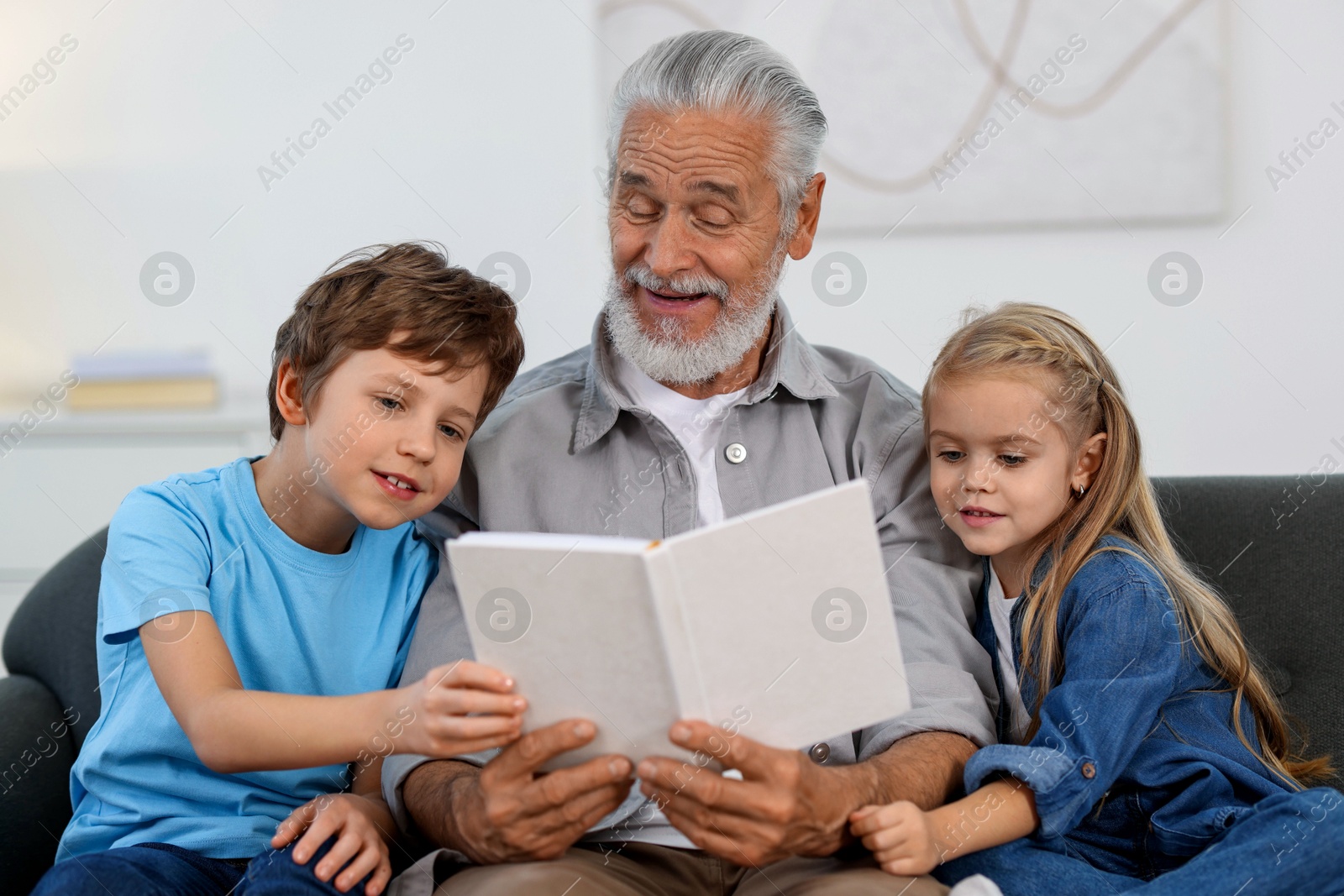 Photo of Grandpa and his grandkids reading book together on sofa at home