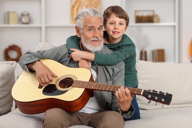 Photo of Grandpa teaching his grandson to play guitar on sofa at home