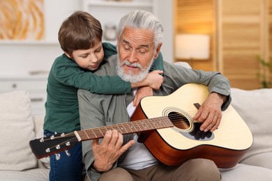 Photo of Grandpa teaching his grandson to play guitar on sofa at home