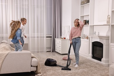 Photo of Tired housewife vacuuming floor while her kids playing in room