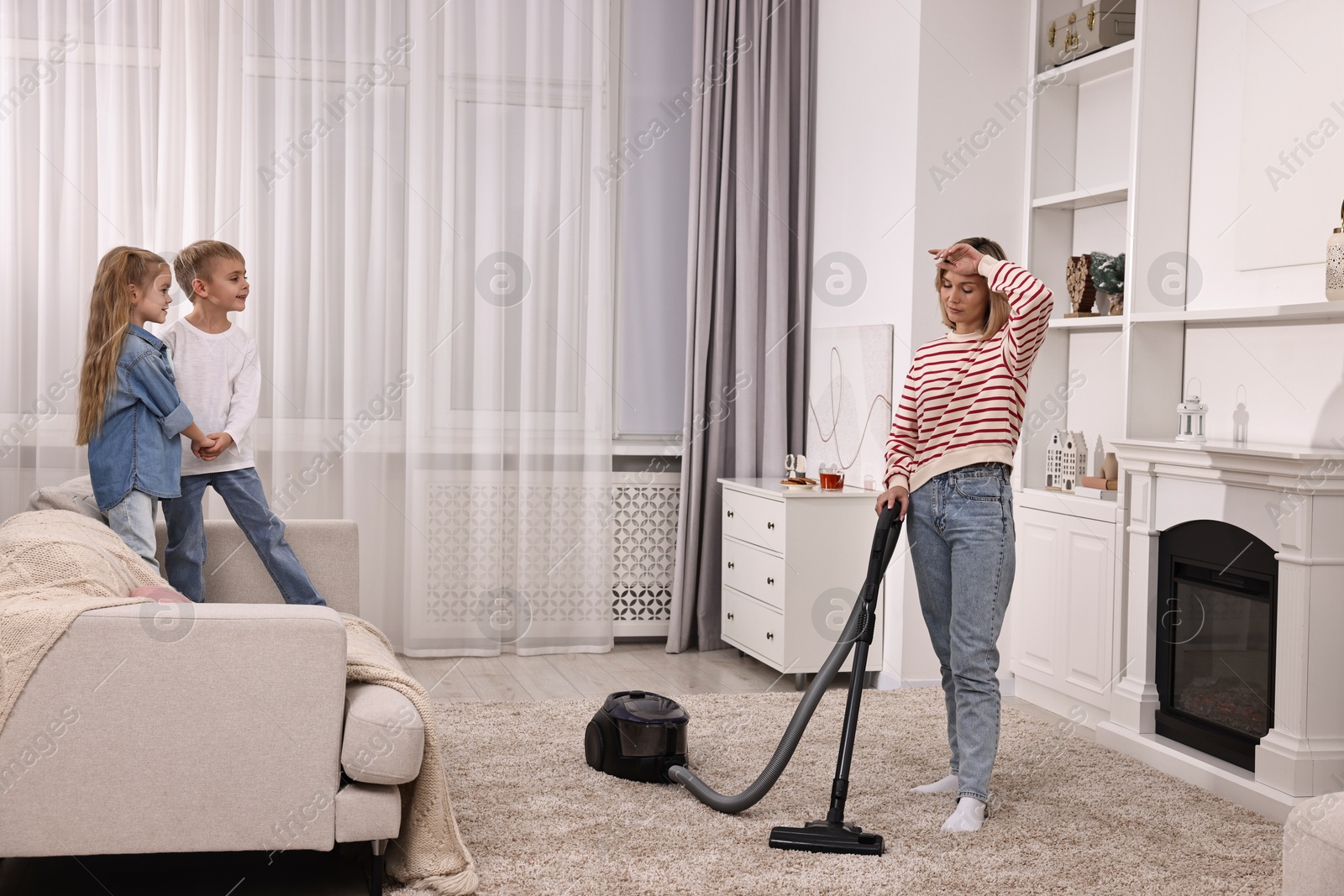 Photo of Tired housewife vacuuming floor while her kids playing in room