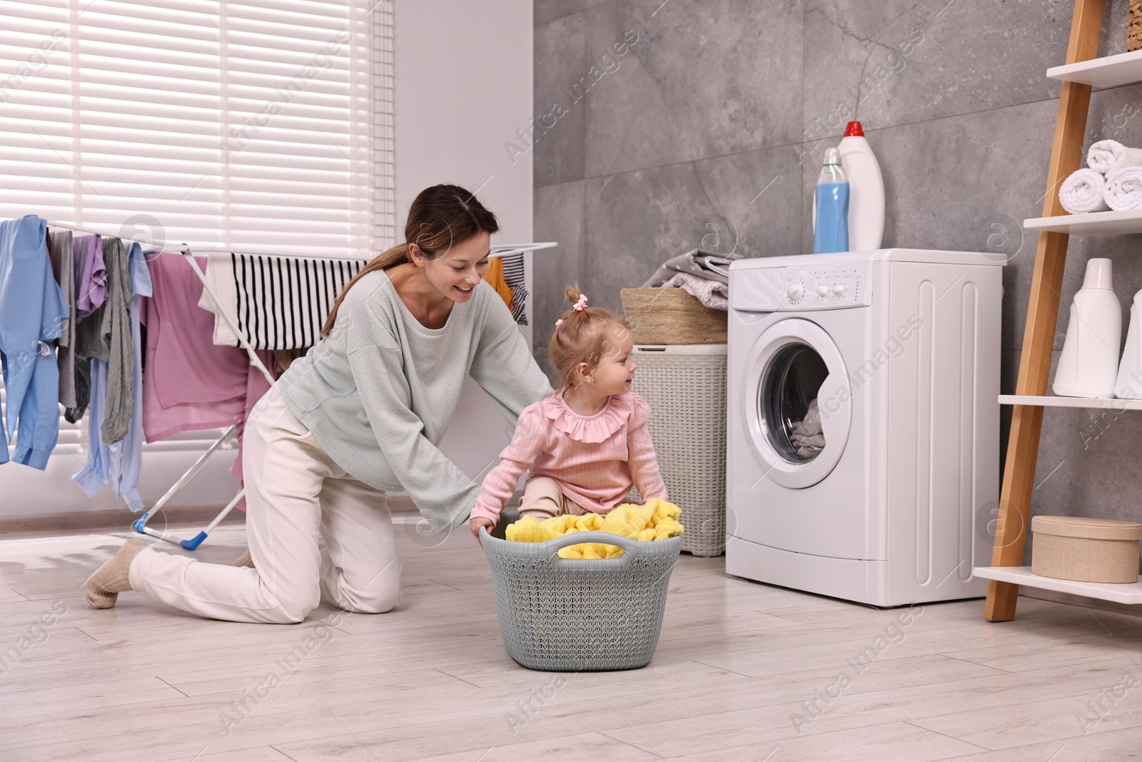 Photo of Smiling housewife and her little daughter having fun with basket of laundry at home