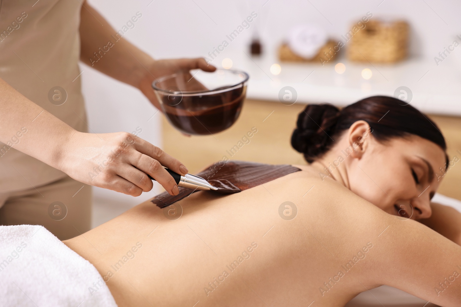 Photo of Chocolate body wrap. Spa worker applying mask onto woman's back in salon, closeup