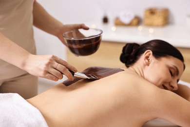 Chocolate body wrap. Spa worker applying mask onto woman's back in salon, closeup