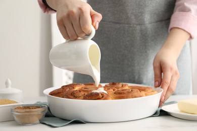 Photo of Woman adding frosting onto freshly baked cinnamon rolls at white table indoors, closeup