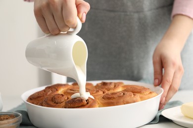 Photo of Woman adding frosting onto freshly baked cinnamon rolls at table indoors, closeup