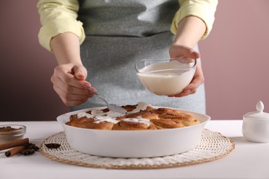 Photo of Woman spreading frosting onto freshly baked cinnamon rolls at white table against pink background, closeup