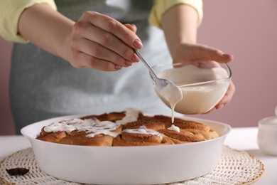 Photo of Woman spreading frosting onto freshly baked cinnamon rolls at white table against pink background, closeup