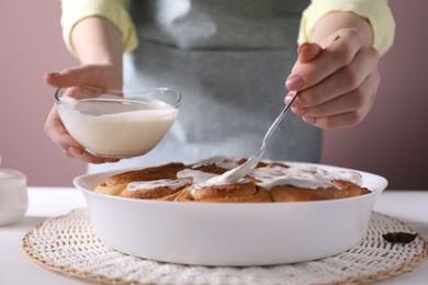 Photo of Woman spreading frosting onto freshly baked cinnamon rolls at white table against pink background, closeup