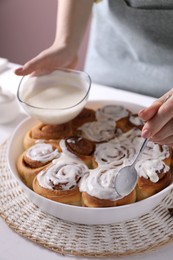 Photo of Woman spreading frosting onto freshly baked cinnamon rolls at white table against pink background, closeup