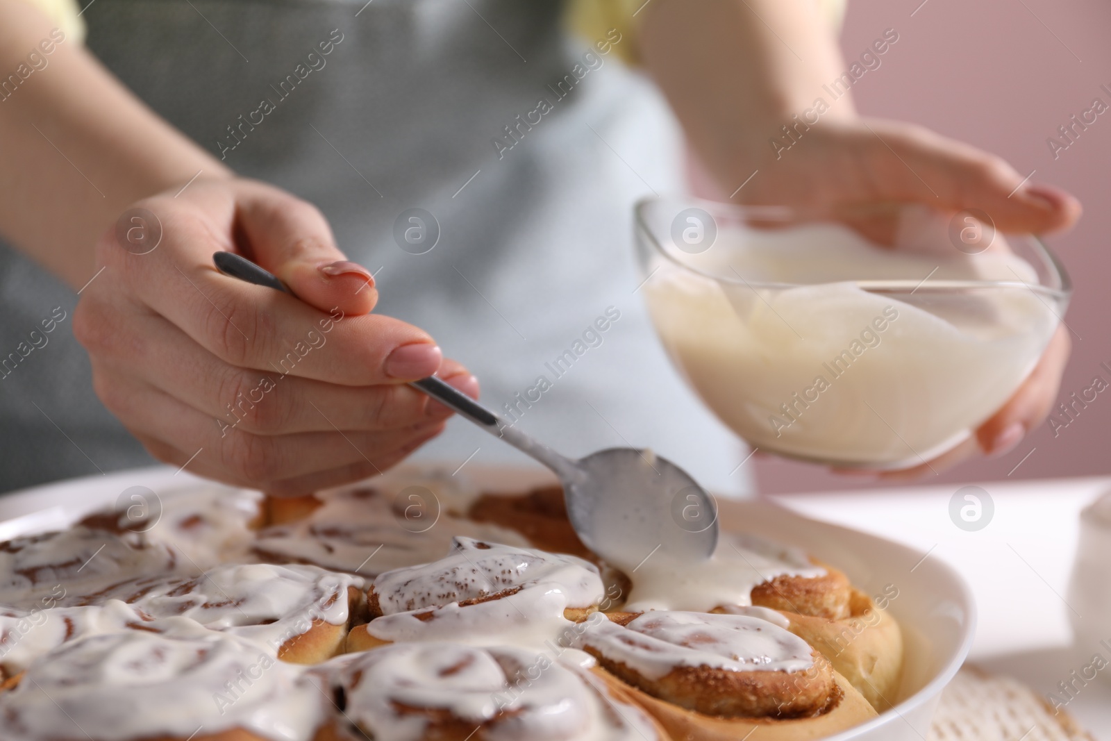 Photo of Woman spreading frosting onto freshly baked cinnamon rolls at white table against pink background, closeup