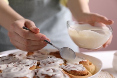 Photo of Woman spreading frosting onto freshly baked cinnamon rolls at white table against pink background, closeup